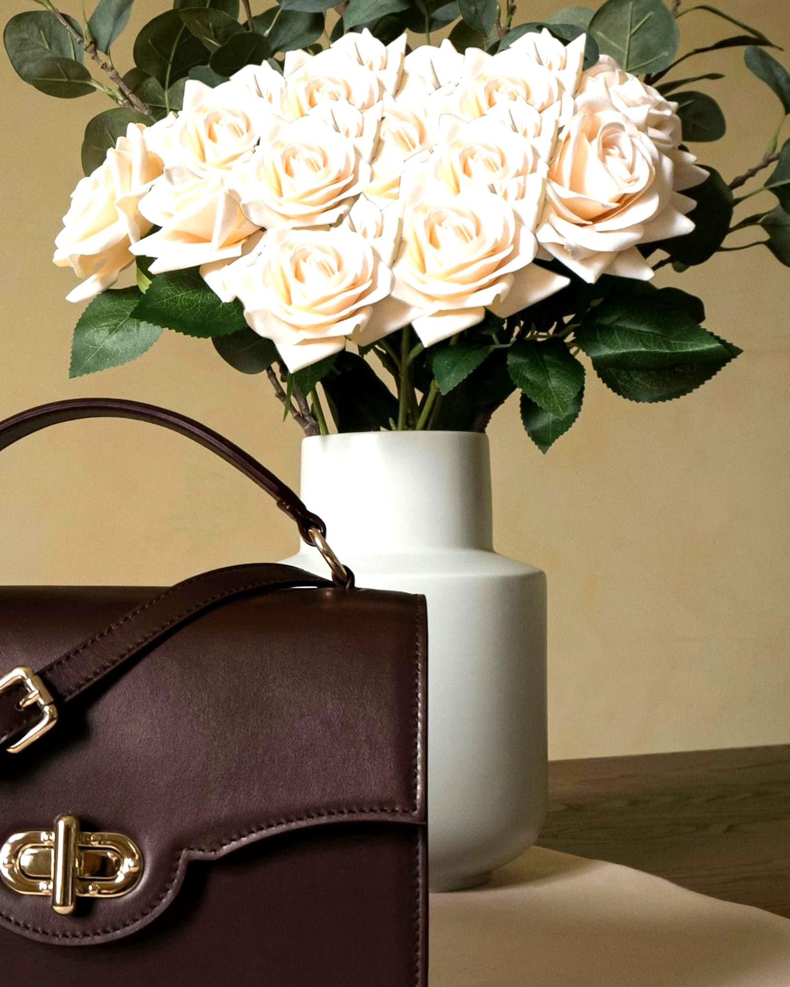Oxblood leather flap bag with a gold twist lock, beside cream roses — detail view