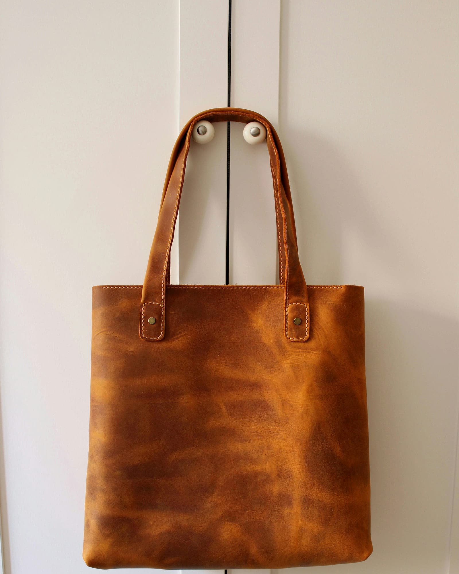 Tan pull-up leather tote with slim handles hanging against a white door