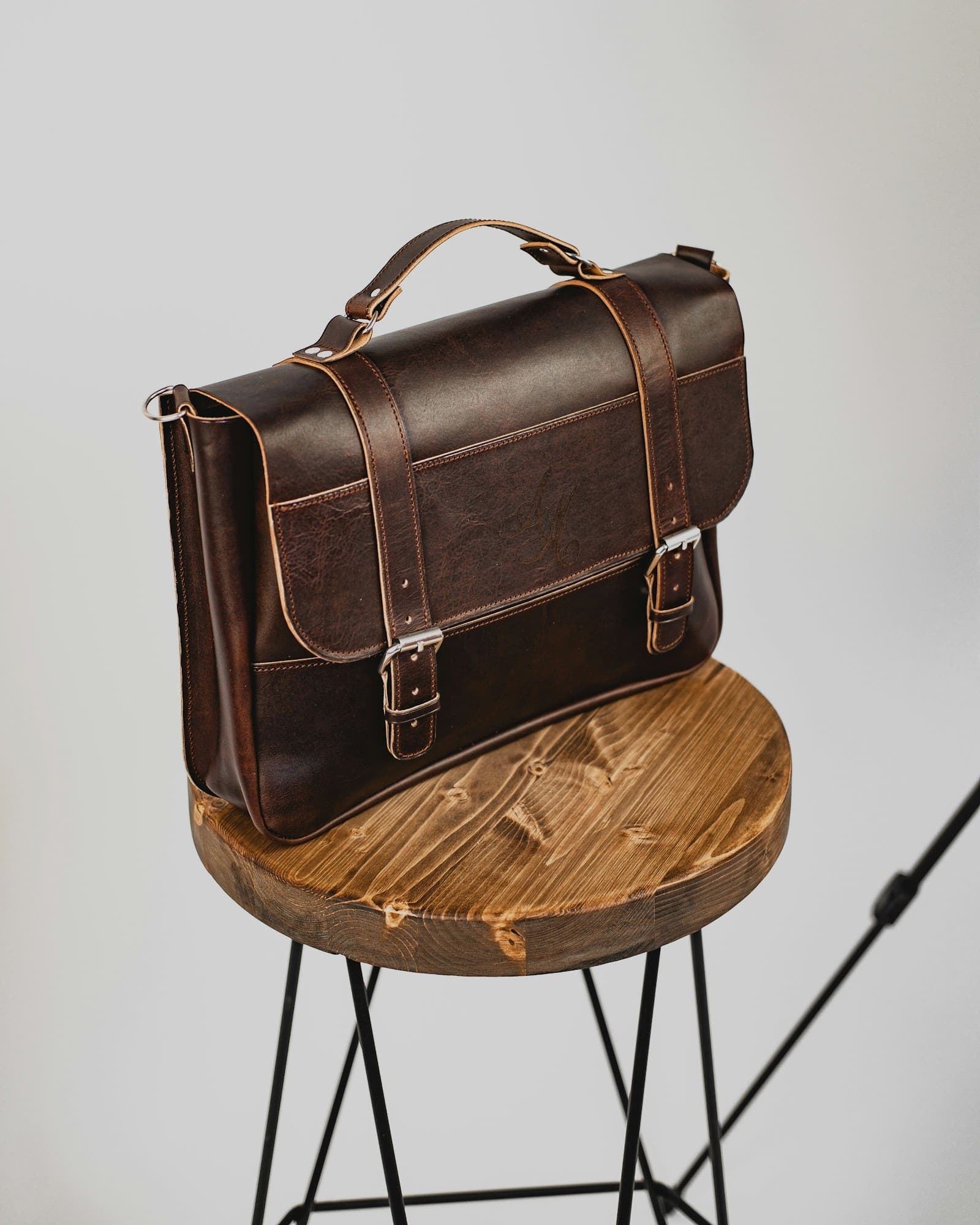 Dark brown leather satchel with buckled straps on a round wooden stool