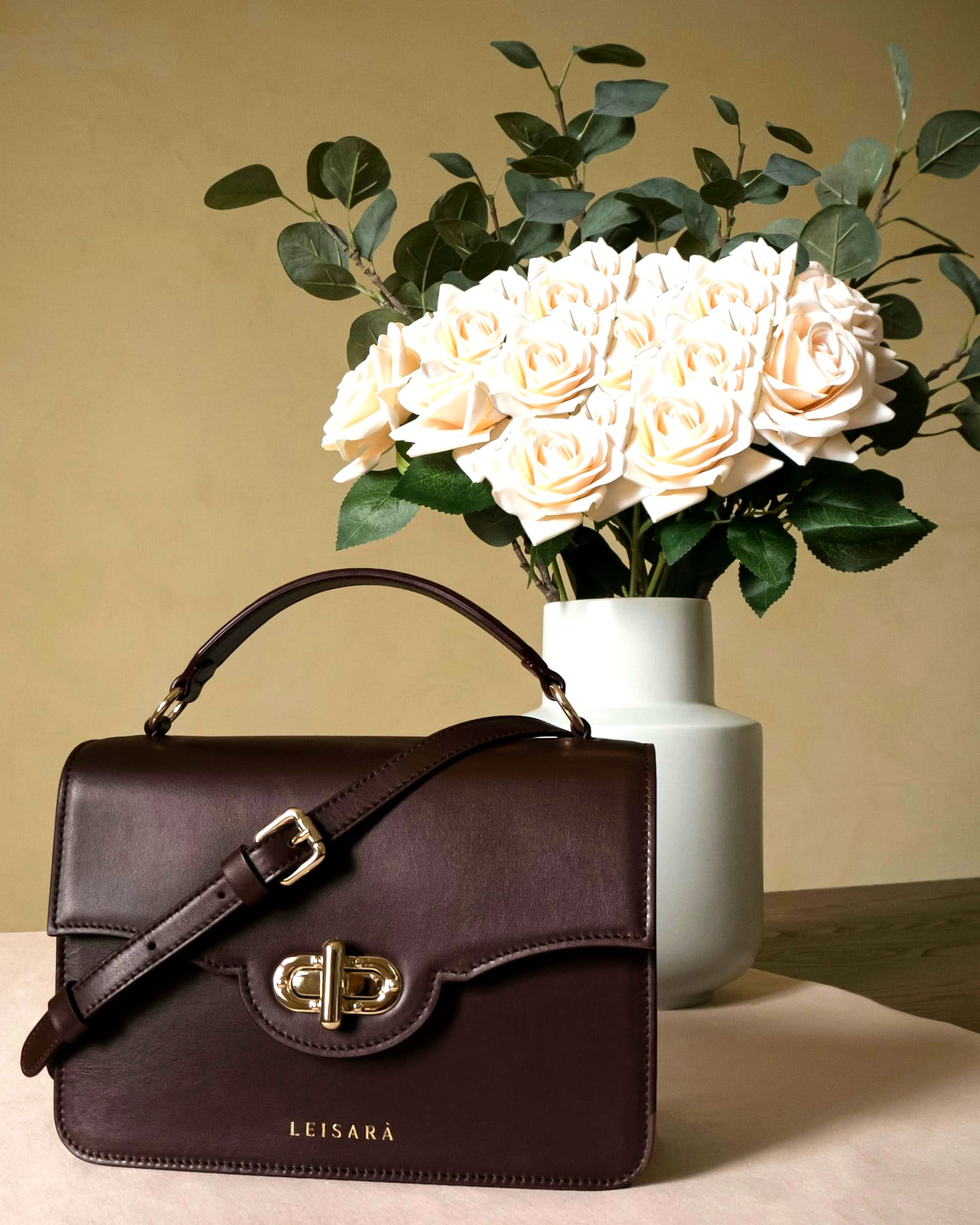 Oxblood leather flap bag with a gold twist lock, beside cream roses