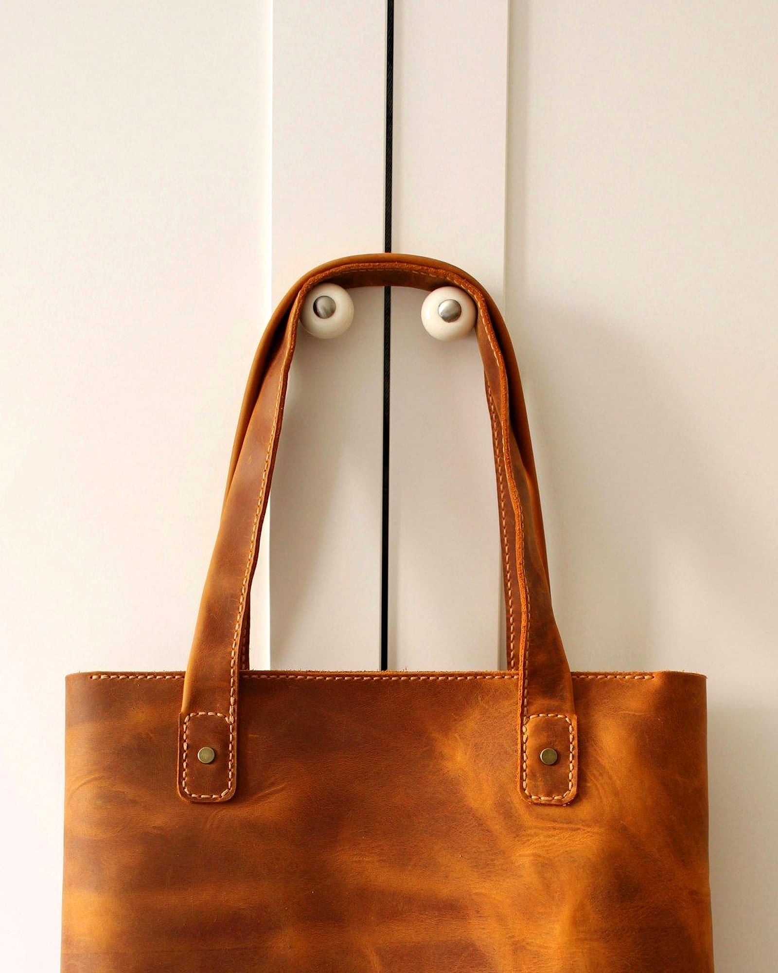 Tan pull-up leather tote with slim handles hanging against a white door — detail view