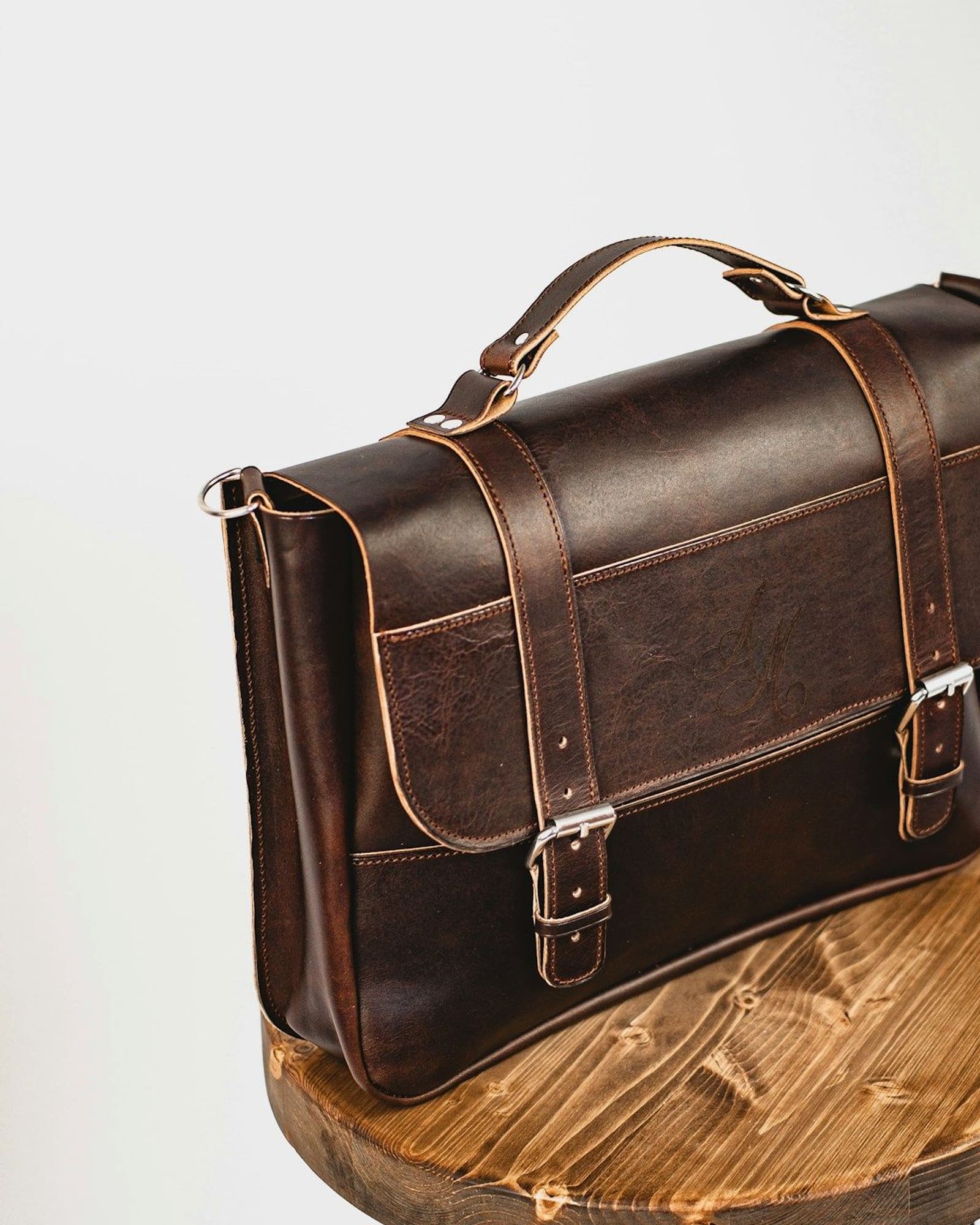 Dark brown leather satchel with buckled straps on a round wooden stool — detail view