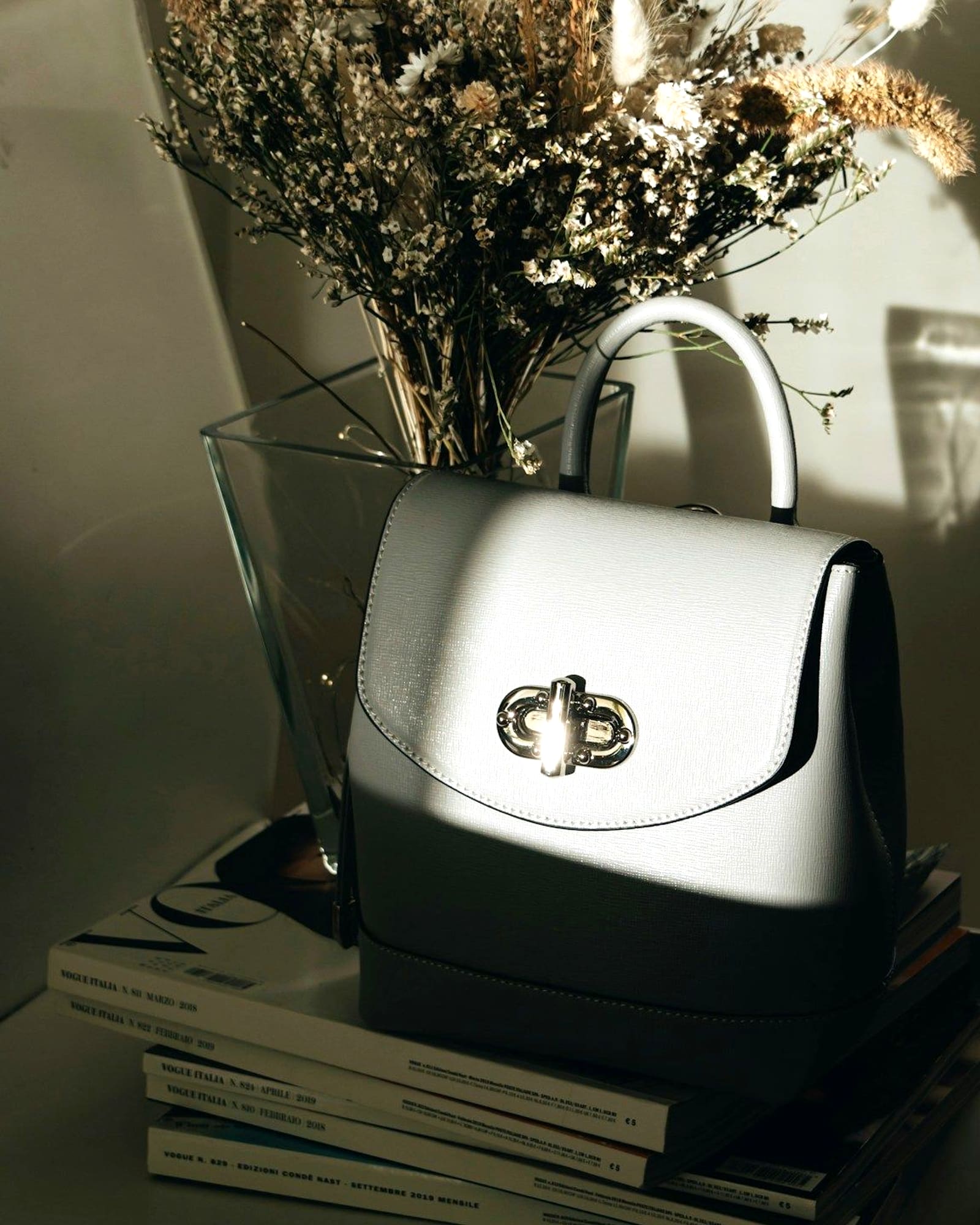 Cream top-handle bag with a silver turn lock, on books beside dried flowers — detail view