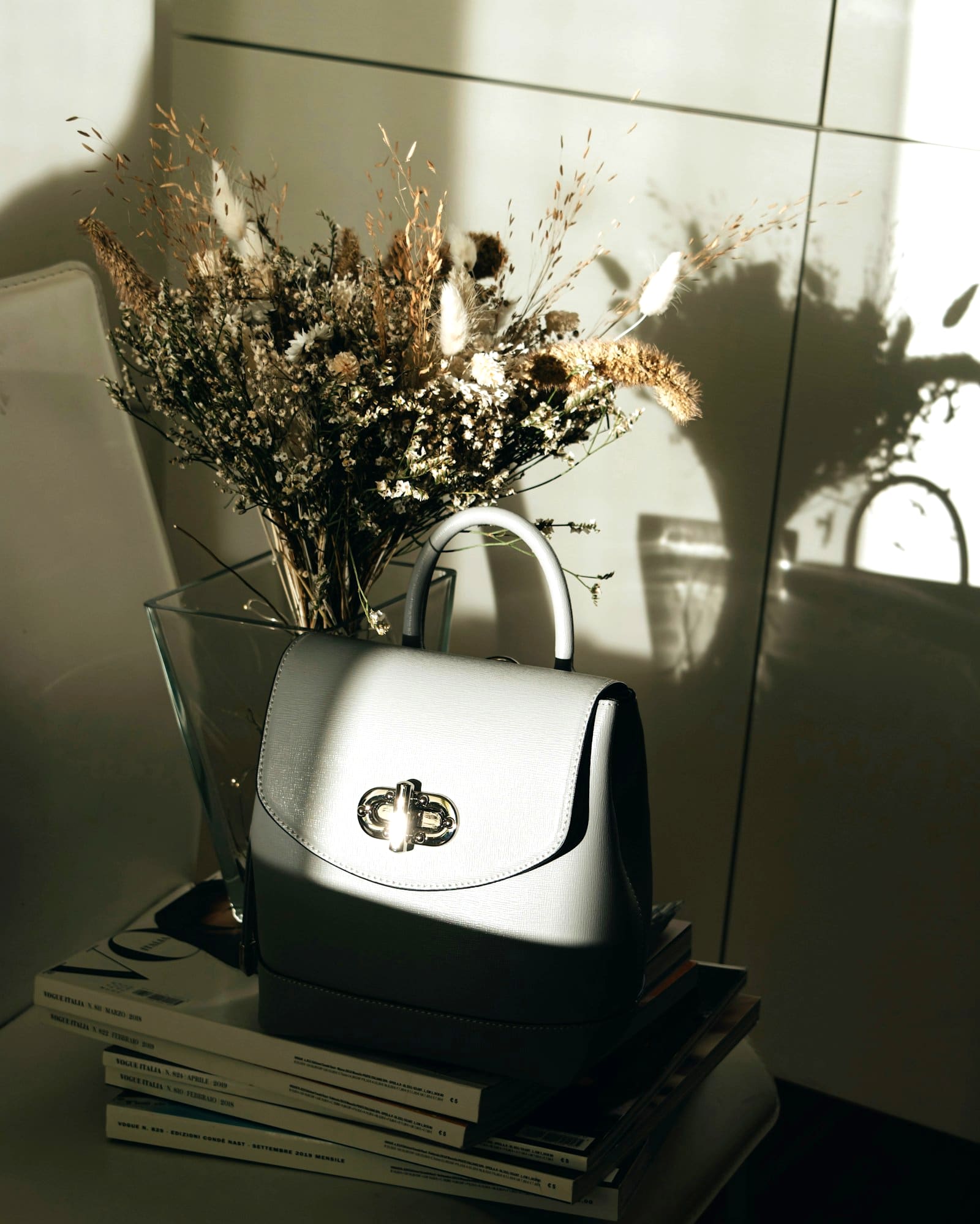Cream top-handle bag with a silver turn lock, on books beside dried flowers