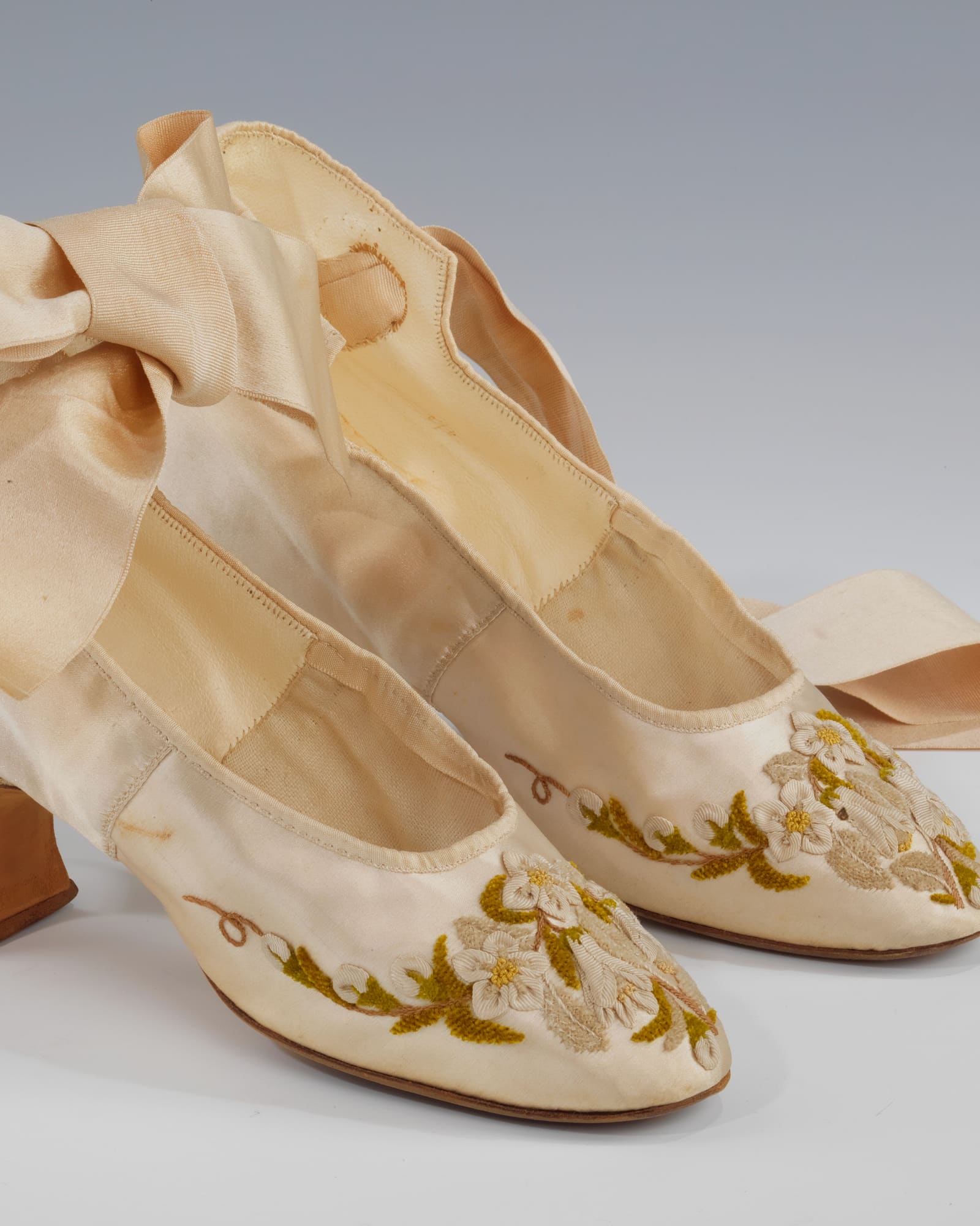 Pair of ivory satin slippers with ribbon ties, grey background — detail view